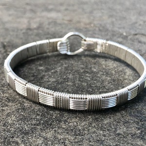 May include: A silver wire-wrapped bracelet. The bracelet has a woven design and a hook clasp. The metal has a bright, reflective finish. This is a piece of jewellery.