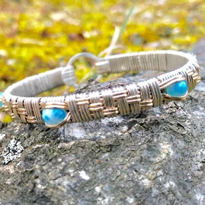Puede incluir: Una pulsera de alambre plateado y dorado con detalles de piedras preciosas azules. La pulsera presenta un diseño tejido con pequeñas piedras preciosas redondas de color azul claro, espaciadas uniformemente alrededor de la banda. El cierre es un simple cierre de gancho y ojo.