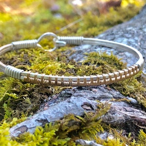 May include: A silver and gold wire-wrapped bracelet. The bracelet has a hook closure and a central section wrapped with gold-coloured wire. The bracelet is resting on a moss-covered log.