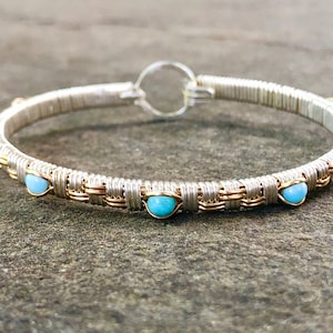 Könnte beinhalten: Silberfarbenes, drahtgewickeltes Armreif-Armband mit goldfarbenen Akzenten und hellblauen Edelsteinperlen. Das Armband hat einen kreisförmigen Verschluss und liegt auf einer strukturierten grauen Oberfläche.