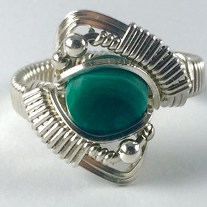 Può includere: Anello artigianale in filo d'argento con una vibrante pietra preziosa verde smeraldo di forma ovale. Il design dell'anello incorpora un'intricata lavorazione a filo, con un focus su motivi geometrici e piccole perle d'argento. La pietra preziosa è il punto focale.