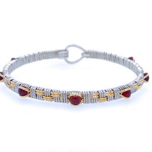 Könnte beinhalten: Ein silber- und goldfarbenes Armband mit roten Edelsteinen. Das Armband hat ein gewebtes Design mit abwechselnd silbernen und goldenen Metallsträngen. Rote Edelsteine sind gleichmäßig verteilt. Der Verschluss ist ein einfacher Haken und Öse.