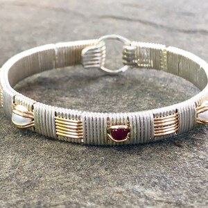 May include: Silver wire-wrapped bracelet with gold detailing. The bracelet has small, round, white pearls and oval, deep red gemstones. The clasp is a simple silver ring. The design of the bracelet is intricate and detailed.