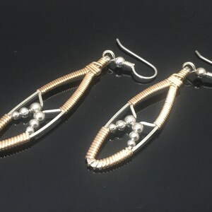 May include: A pair of handcrafted earrings with a teardrop design. The earrings have a silver frame, gold-coloured wire wrap, and a cluster of silver beads. The ear wires are silver.