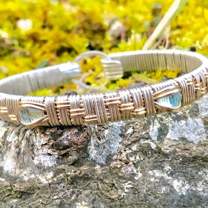 May include: A handmade bracelet with a silver and gold woven pattern. The bracelet incorporates three light blue, teardrop-shaped gemstones. The clasp is silver. The bracelet is shown on a textured, natural surface.