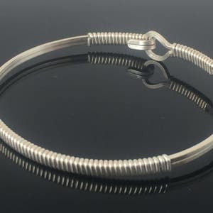 May include: A silver bangle bracelet with a hook clasp. The bracelet has a smooth, rounded band with coiled wire detailing at each end and near the clasp. The hook clasp is also silver-toned.