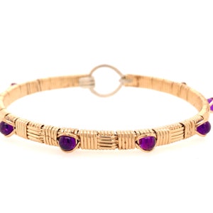 May include: A gold-coloured bangle bracelet with woven wire detailing. The bracelet features several round, deep purple gemstones evenly spaced around the band. The clasp is a simple silver-coloured loop.