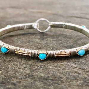 May include: Silver wire-wrapped bracelet with gold accents and turquoise beads. The bracelet has a circular design with a clasp. The beads are evenly spaced around the bracelet. The bracelet is approximately 18 cm in circumference.