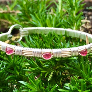 May include: A silver and gold wire-wrapped bracelet with pink gemstones. The bracelet features a series of pink, teardrop-shaped stones set along the band. The wire is intricately woven, creating a delicate design. The bracelet is set against a backdrop of green foliage.