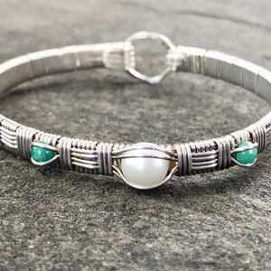 May include: A silver wire-wrapped bracelet featuring a central white pearl and two green beads. The bracelet has a clasp closure and is made of silver-coloured wire. The design is intricate with a woven pattern around the bracelet's circumference.