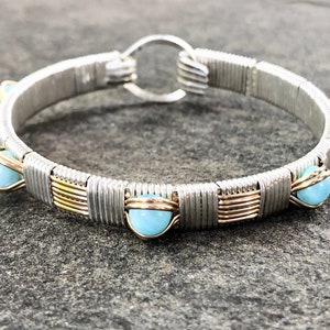 Può includere: Un braccialetto argentato avvolto in filo metallico con accenti di pietre preziose blu chiaro. Il braccialetto presenta un design circolare con chiusura a fermaglio. Il design incorpora accenti di filo dorato, aggiungendo contrasto allo schema di colori argento e blu.