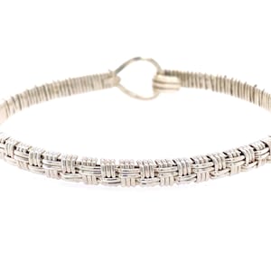 Könnte beinhalten: Silberfarbenes Armband mit einem gewebten Design in der Mitte. Das Armband hat einen Haken- und Ösenverschluss. Der gewebte Bereich ist etwa 1,3 cm breit. Auf weißem Hintergrund.