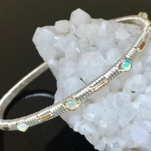 May include: Silver bangle bracelet featuring small, oval opals set along the band. The bracelet has a textured, wrapped design with gold accents. The opals have a milky, iridescent appearance. The bracelet is displayed against a white background.