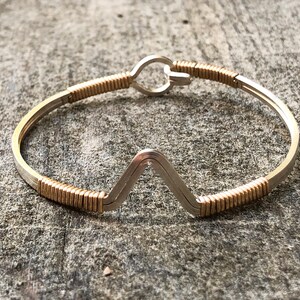May include: A silver and gold-toned metal bracelet. The bracelet has a V-shaped design in the centre and is accented with gold-coloured wire wrapping. The clasp is a simple hook and eye closure. The jewellery is displayed on a textured grey surface.
