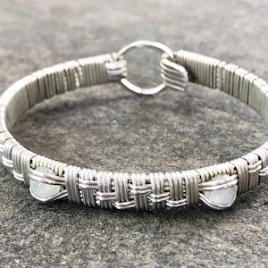 Könnte beinhalten: Ein silberfarbenes, drahtgewickeltes Armband mit einem gewebten Design. Das Armband hat kleine, runde, hellfarbige Steine und einen kreisförmigen Verschluss. Der Schmuck wird auf einer grauen Oberfläche präsentiert.