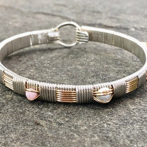 May include: Silver wire-wrapped bangle bracelet with gold accents and small pink and white beads. The bracelet has a circular clasp. The jewellery is displayed on a grey surface. Handmade.