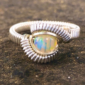 May include: A silver wire-wrapped ring featuring a round, iridescent opal gemstone. The ring's band and setting are crafted from coiled silver wire, creating a textured and artistic design. The opal displays a play of colors.