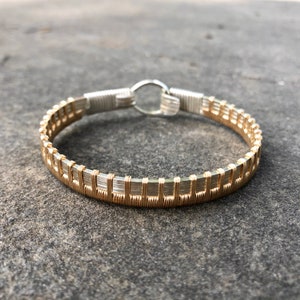 May include: A gold and silver wire-wrapped bracelet. The bracelet has a rectangular pattern in gold wire, with silver wire accents and a silver clasp. The jewellery is displayed on a grey surface.