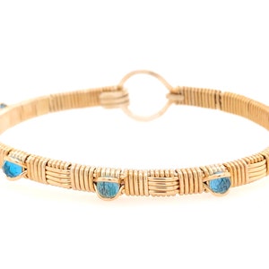 May include: A gold-coloured wire-wrapped bracelet with a circular clasp. The bracelet has several light blue gemstones evenly spaced around the band. The gemstones contrast with the gold wire.