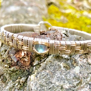 May include: A silver and gold woven bracelet with three oval-shaped, iridescent gemstones. The bracelet features a woven design with silver and gold wire, and the gemstones have a blue and green colour. The bracelet is set against a natural, textured background.