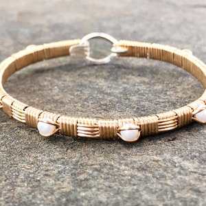 May include: A gold wire wrapped bangle bracelet with white pearl accents. The bracelet is made of fine wire and has a delicate, feminine look.