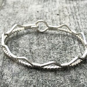 May include: A silver wire-wrapped ring with a distinctive, undulating design. The ring is crafted from thin, shiny metal and features a series of connected geometric shapes. The ring is displayed on a grey, textured surface.