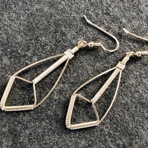 May include: A pair of silver wire earrings. Each earring has a teardrop shape with a geometric diamond at the base. The wire is tightly wound in sections, creating a textured effect. The earrings have a simple hook closure.