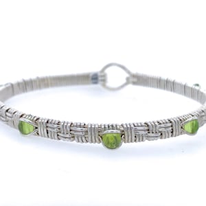 Könnte beinhalten: Ein silberfarbenes Armreif-Armband mit geflochtenen Drahtdetails und mehreren hellgrünen Peridot-Edelsteinen. Das Armband hat einen Haken- und Ösenverschluss. Die Edelsteine sind gleichmäßig verteilt.