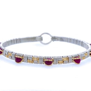 May include: A silver and gold wire-wrapped bracelet with vibrant red gemstones. The bracelet features a woven design with alternating silver and gold wire, and the gemstones are evenly spaced around the band.