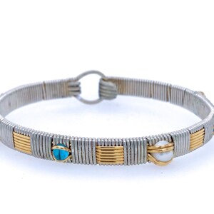 Può includere: Bracciale in filo d'argento e oro con accenti di pietre preziose turchesi e bianche. Il bracciale presenta un design circolare con chiusura a fermaglio. Le pietre preziose sono uniformemente distanziate.