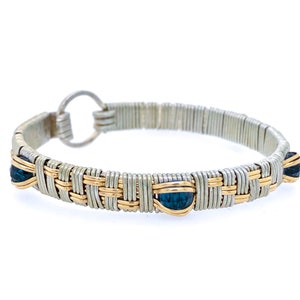 May include: A silver and gold-toned wire-wrapped bracelet with three dark blue gemstone accents. The bracelet features a woven design with a circular clasp. The gemstones are teardrop-shaped.