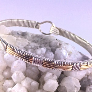 May include: A silver and gold-toned wire-wrapped bracelet. The bracelet features a woven design with alternating silver and gold-colored wire sections. The clasp is a simple hook and eye closure. The jewelry is displayed on a white, crystalline surface.