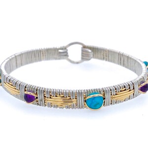 Puede incluir: Un brazalete de alambre de plata y oro. La pulsera presenta piedras preciosas de turquesa y amatista alternadas. La pulsera tiene un cierre de gancho y ojo. La joya está sobre un fondo blanco.