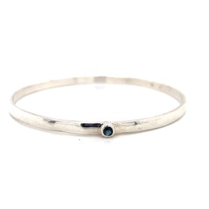 Può includere: Un braccialetto rigido in argento con una singola pietra preziosa blu incastonata al centro.