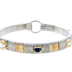 May include: Silver and gold-toned wire-wrapped bracelet. The bracelet features alternating sections of silver wire and gold wire. It is adorned with pearls and dark blue gemstones. The bracelet has a circular clasp.
