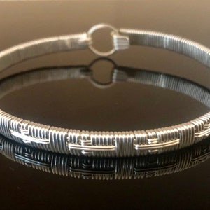 May include: A silver wire bangle bracelet with a woven design. The bracelet is open and has a small loop for fastening.