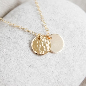 May include: A delicate gold necklace featuring two pendant charms. One charm has a hammered texture, while the other is smooth. The necklace is displayed on a light gray stone, with a soft, neutral background.