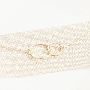 May include: A gold necklace with two interlinked circles. The circles are joined by a fine chain, creating a minimalist and elegant design. The necklace is displayed on a neutral-coloured fabric, highlighting its simple beauty.