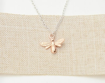 May include: A rose gold bee pendant necklace with a delicate silver chain. The bee pendant has detailed wings and body, and is displayed on a neutral linen background. The necklace is a piece of jewelry.