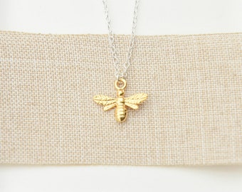 May include: A gold-colored bee pendant necklace with a silver chain. The bee pendant has detailed wings and body segments. The necklace is displayed on a beige fabric background.