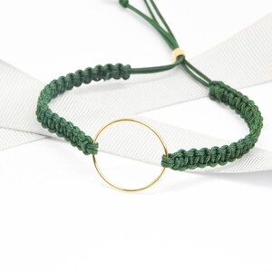 May include: A green braided bracelet with a gold-colored circular charm. The bracelet is adjustable with a sliding knot closure. The background is white with a white ribbon.