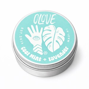 May include: A round, silver tin with a turquoise lid featuring the word "OLOVE" in white script. The lid also displays a white hand with an eye, a monstera leaf, and the text "Cool Mint + lavender". The text "EST. 2014" and "MAUI, HI" are also visible.