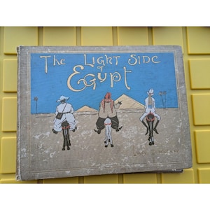 Pode incluir: A capa de um livro antigo intitulado "The Light Side of Egypt" com uma ilustração de três pessoas montando burros em frente a pirâmides.