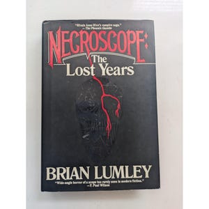 Puede incluir: Un libro de tapa dura de Brian Lumley, titulado "Necroscope: The Lost Years". La portada negra presenta un diseño de calavera con detalles rojos. Incluye citas de The Phoenix Gazette y F. Paul Wilson.