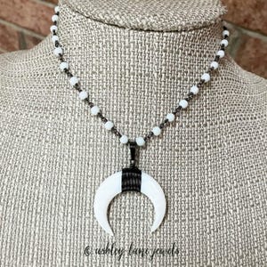 May include: A white crescent moon pendant necklace with a black wire wrap and a beaded chain. The necklace is displayed on a mannequin. The text "© ashley lane jewels" is visible at the bottom.