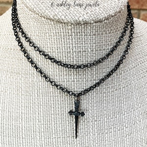 May include: Black layered necklace with a dagger pendant. The necklace features two strands of linked chain. The dagger pendant has a cross-shaped hilt and a pointed blade. The text "ashley lane jewels" is in the upper right corner.