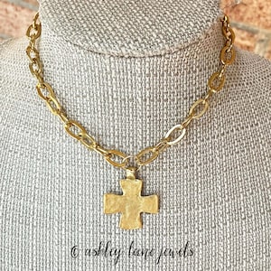 May include: Gold-toned necklace with a large cross pendant. The necklace features a chunky chain design and a textured cross. The text "© ashley lane jewels" is visible at the bottom of the image.