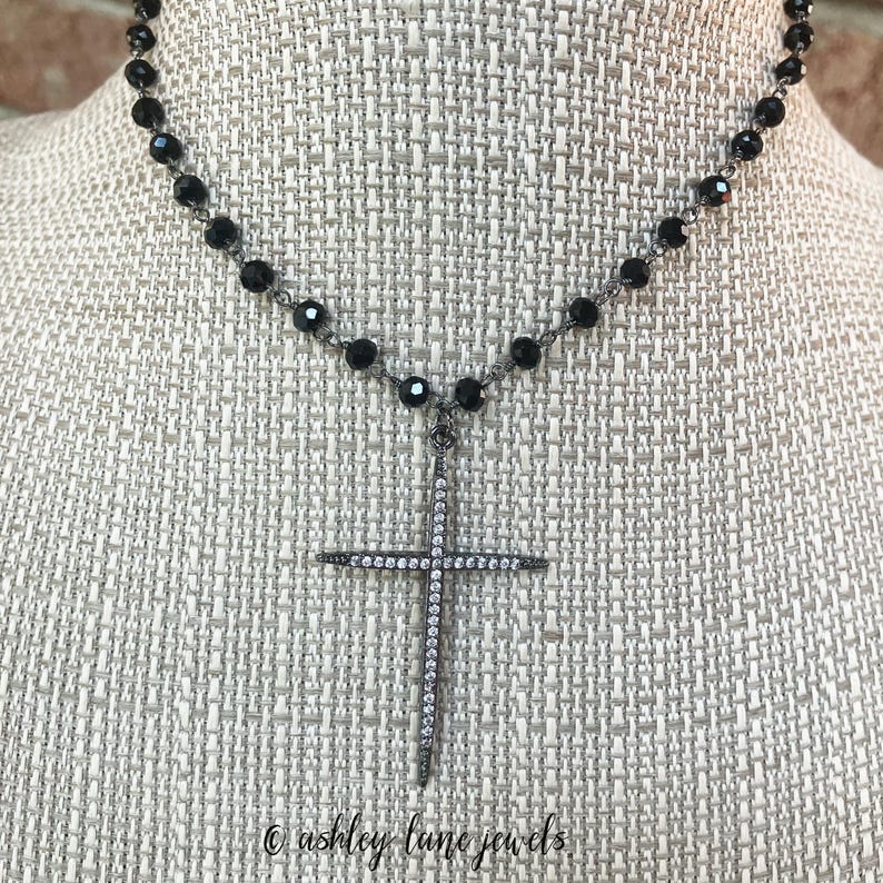 May include: A black beaded necklace with a cross pendant. The cross is encrusted with small, clear crystals. The necklace is displayed on a neutral-colored surface. The text "&copy; ashley lane jewels" is at the bottom.