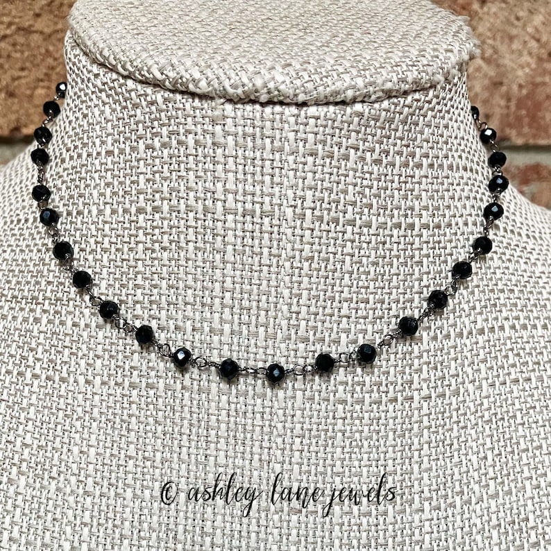 May include: A delicate necklace featuring small, faceted black beads spaced evenly along a thin, dark metal chain. The necklace is displayed on a textured fabric form, with the text ashley lane jewels printed in black script at the bottom.
