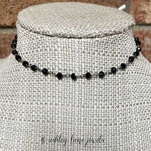May include: A delicate choker necklace featuring small, faceted black beads strung along a thin, dark metallic chain. The necklace is displayed on a textured, light-colored fabric form. The text ashley lane jewels is visible at the bottom.